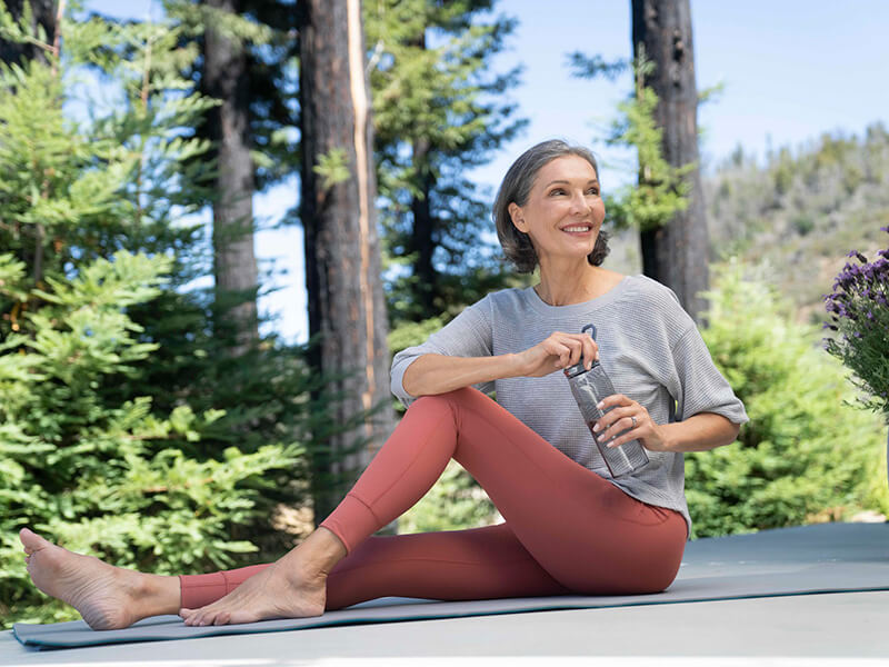 An older woman is stretching after a yoga session, holding a bottle of water in her hands.