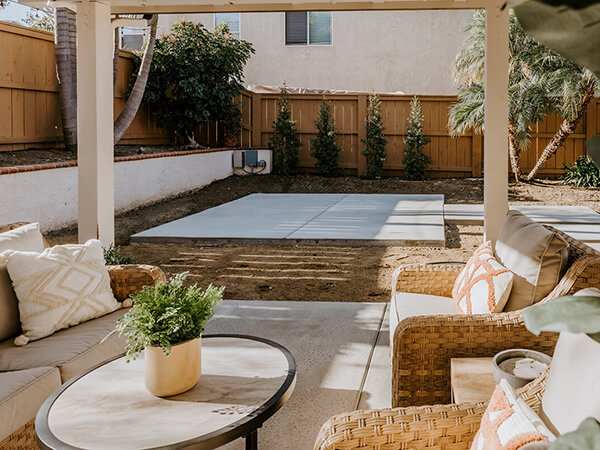  A spacious backyard with a concrete patio and a covered seating area. The patio features comfortable wicker furniture, potted plants, and a view of a lush green lawn.